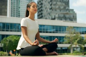 37752845 - young stressed hispanic business woman doing yoga outside office building, sitting in lotus position with hands on knees in the street. concept of long working hours and need of stress free break